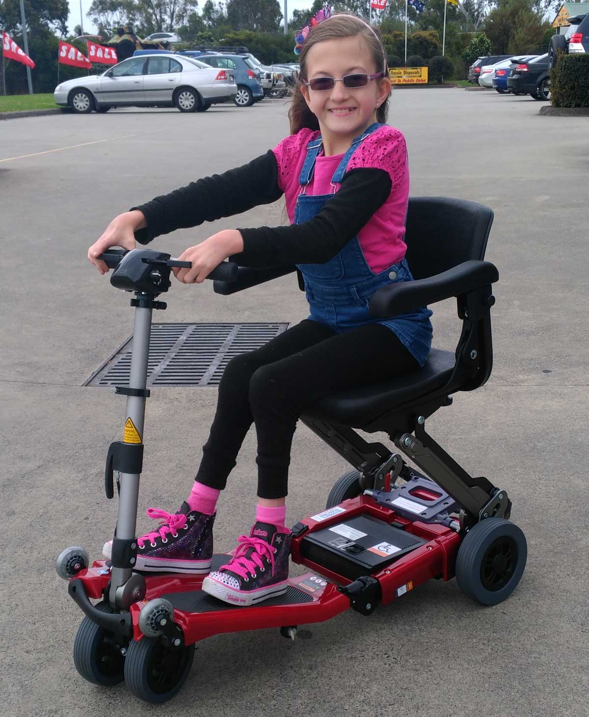 Young girl sitting on a red, sleek, mobility scooter in a parking lot.